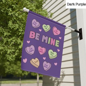 May include: A dark purple garden flag with the words "BE MINE" in pink and green. The flag features heart-shaped candies with phrases like "Free Press," "Civil Rights," and "Social Justice."