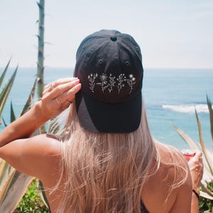 May include: A black baseball cap with white embroidered flowers. The cap has a curved brim and an adjustable strap.