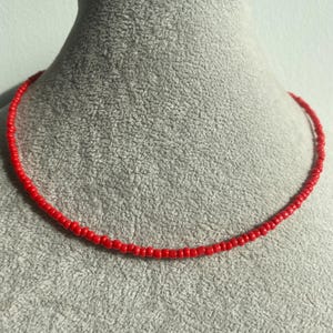 Puede incluir: Un collar de cuentas rojo brillante exhibido en un maniquí gris. El collar está compuesto por pequeñas cuentas espaciadas uniformemente, creando un diseño circular continuo. El color rojo brillante de las cuentas contrasta con el fondo neutral, resaltando la joya.