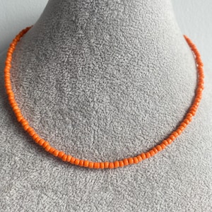 May include: A simple orange beaded necklace on a grey mannequin. The necklace is made of small, round beads.