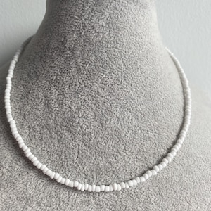 Può includere: Una delicata collana di perline bianche esposta su un manichino grigio. La collana è composta da piccole perline bianche rotonde, creando un design semplice ed elegante. Le perline sono infilate insieme per formare una collana.