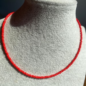 Puede incluir: Un collar de una sola hebra compuesto por pequeñas cuentas rojas uniformes y vibrantes. El collar tiene forma circular y presenta una textura consistente y suave a lo largo de toda la hebra, creando un diseño simple y continuo.