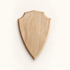 Può includere: Una targa in legno marrone chiaro a forma di scudo araldico. La superficie presenta una venatura naturale del legno con linee verticali che corrono dalla parte superiore alla punta inferiore. I bordi sono smussati, creando un bordo definito.