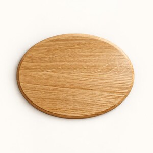 Pode incluir: Uma placa de madeira de formato oval com um padrão de grão de madeira castanho-claro natural. A superfície é lisa com uma textura visível de madeira de carvalho, exibindo linhas horizontais e padrões orgânicos em toda a face plana do objeto.