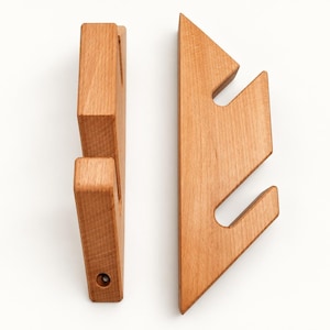 Pode incluir: Dois ganchos de parede de madeira castanho-claro com perfil triangular e entalhes angulares. Cada gancho apresenta um acabamento de grão liso e um orifício de montagem circular na base, concebido para pendurar objetos numa superfície.