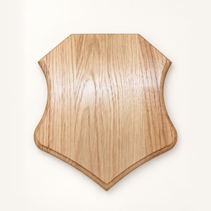 May include: A light brown wooden plaque with a smooth, polished finish and a distinct wood grain pattern. The plaque features a shield-like shape with a flat top, angled upper corners, and curved sides that taper down to a pointed bottom edge.