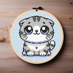 以下が含まれることがあります： 大きな青い目をした、かわいいグレーと白の漫画の猫が描かれたクロスステッチ刺繍フープ。猫は薄い青色の背景に座っています。