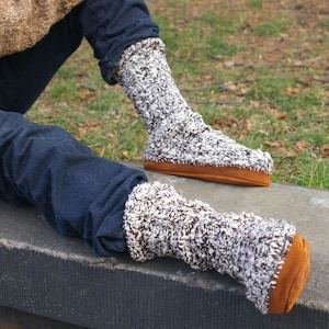 Op de afbeelding: Een paar bruine en witte fuzzy pantoffels met een bruine suède zool. De sokken worden gedragen aan de voeten van iemand.
