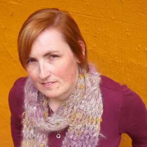 May include: A woman with red hair wearing a maroon shirt and a light purple and beige scarf. The scarf is a chunky knit with a loose weave.