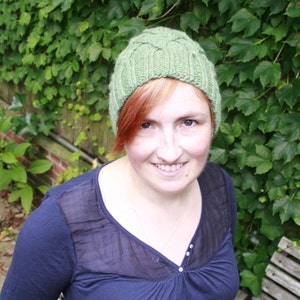 Op de afbeelding: Een vrouw draagt ​​een groene gebreide muts met een kabelbreipatroon. Ze glimlacht en heeft rood haar.