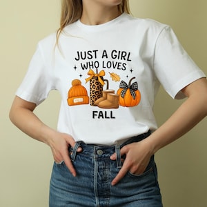 Puede incluir: Camiseta blanca con el texto "JUST A GIRL WHO LOVES FALL" e ilustraciones de temática otoñal. El diseño incluye un gorro naranja, un regalo con estampado de leopardo, botas marrones y una calabaza.