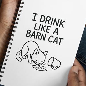May include: A white spiral-bound notebook with the text "I DRINK LIKE A BARN CAT" and a black and white illustration of a cat drinking from a puddle. The notebook is held in a person's hands.