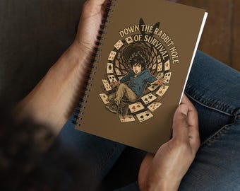 May include: A brown spiral-bound notebook with the text "DOWN THE RABBIT HOLE OF SURVIVAL" and an illustration of a person falling through playing cards. The notebook is held in someone's hands. The cover is brown.
