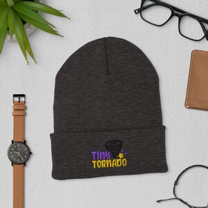 Op de afbeelding: Een donkergrijze beanie met een geborduurd ontwerp in paars en geel met de tekst "Tiny Tornado".