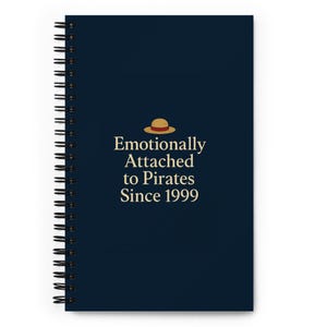 May include: A spiral-bound notebook with a navy blue cover. The cover features the text "Emotionally Attached to Pirates Since 1999" in gold lettering, along with a small illustration of a straw hat. The notebook has a black spiral binding.