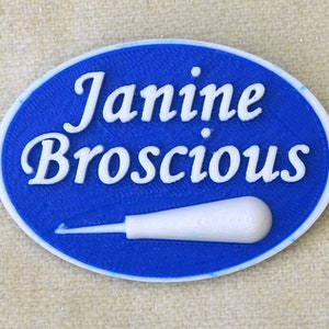 May include: A blue oval-shaped name tag with white text that reads "Janine Broscious". A small white tool is in the bottom center of the tag.
