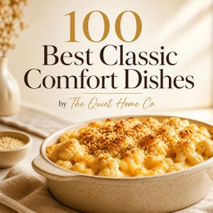 Könnte beinhalten: Ein Kochbuchcover mit dem Titel "100 Best Classic Comfort Dishes" in gold- und braunfarbenem Text. Das Bild zeigt ein gebackenes Gericht mit Blumenkohl und einer Krümelkruste. Eine kleine Schüssel mit Getreide und eine Vase befinden sich im Hintergrund.