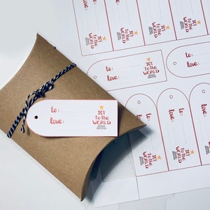 May include: A kraft paper gift box tied with black and white twine, with a white gift tag that reads "to:" and "love:" in red script. The tag also features the text "Joy to the World" and a star graphic. Additional gift tags are visible in the background.