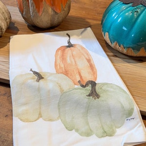 May include: A white kitchen towel with a watercolor painting of three pumpkins. The pumpkins are orange, white, and green. The towel has the text "Art by Ranger" in the bottom right corner.