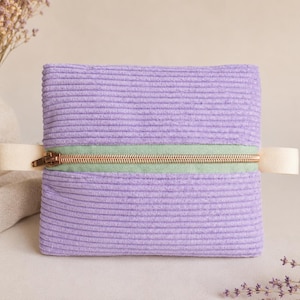 Puede incluir: Un estuche rectangular de pana color lavanda con una franja horizontal de tela verde claro en el centro. Una cremallera metálica dorada recorre el medio de la franja verde, flanqueada por dos lengüetas de tela color blanco hueso.
