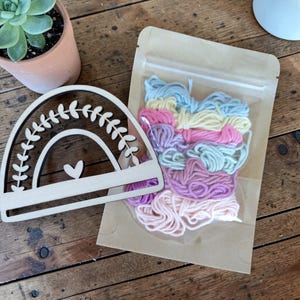 Könnte beinhalten: Ein hölzernes Regenbogen-Bastelstück mit einem Herz-Ausschnitt und Blattdetails, neben einer transparenten Tüte mit farbenfrohem Stickgarn in Rosa-, Blau- und Gelbtönen. Die Artikel befinden sich auf einer Holzoberfläche.