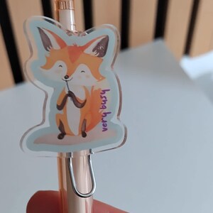 Puede incluir: Un bolígrafo oro rosa con un dije en forma de zorro. El dije presenta un zorro naranja con vientre blanco y orejas marrones, sobre un fondo azul claro. El texto "very busy" está escrito en morado.