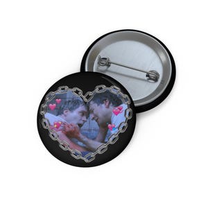 Könnte beinhalten: Ein schwarzer Knopf mit einem herzförmigen Kettenmuster, das zwei sich umarmende Männer zeigt. Das Herz ist mit rosa Herzen gefüllt und der Hintergrund ist schwarz.
