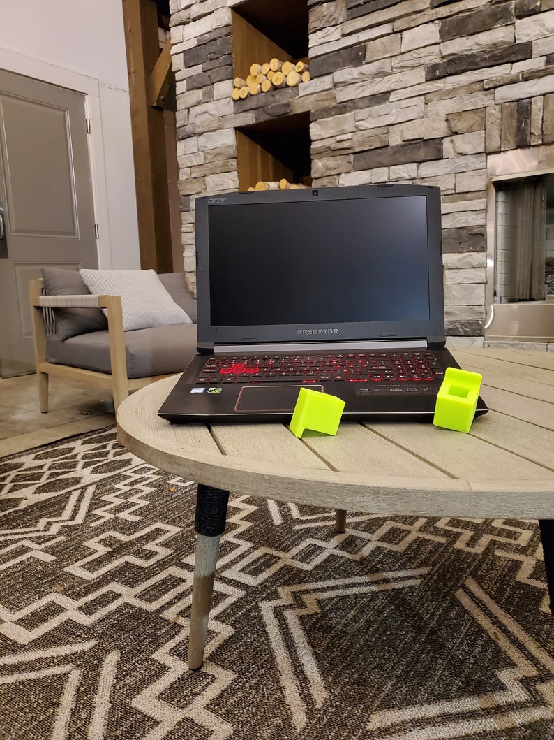 May include: A black Acer Predator laptop computer with a red backlit keyboard is on a wooden table with two yellow blocks. The laptop is in front of a stone fireplace.