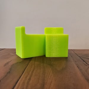 May include: Two lime green 3D printed objects, one is a rectangular block and the other is a rectangular block with a cutout on one side, both are sitting on a wooden surface.