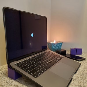 May include: A silver laptop computer with a black case is on a purple stand. The laptop is open and the screen is black with a white Apple logo. A teal candle holder with a lit candle is in the background.