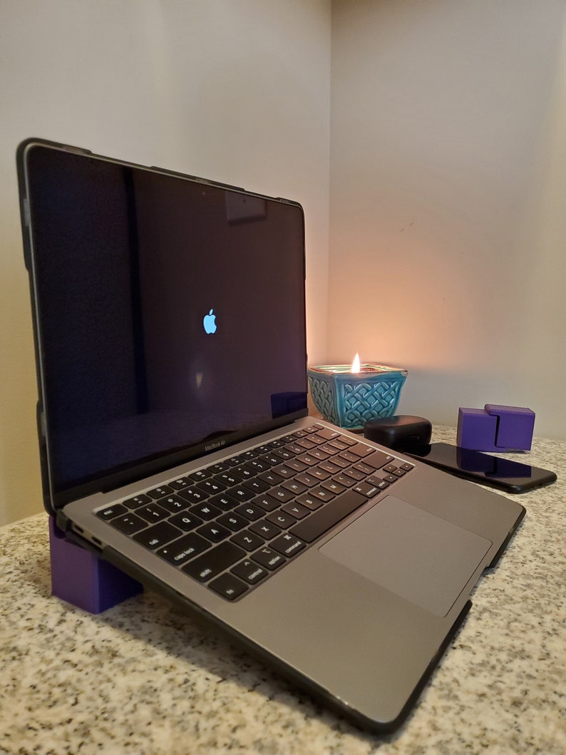 May include: A silver laptop computer with a black case is on a purple stand. The laptop is open and the screen is black with a white Apple logo. A teal candle holder with a lit candle is in the background.