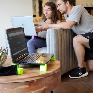 May include: Two people are sitting on a couch, looking at a laptop computer. The laptop is on a table with a wooden top and black legs. The table has four green blocks on the bottom to raise it up. The laptop is open and the screen is showing a website. The person on the left is wearing a blue shirt and the person on the right is wearing a gray shirt.