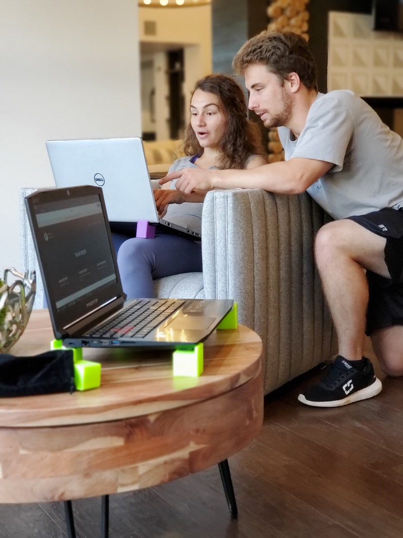 May include: Two people are sitting on a couch, looking at a laptop computer. The laptop is on a table with a wooden top and black legs. The table has four green blocks on the bottom to raise it up. The laptop is open and the screen is showing a website. The person on the left is wearing a blue shirt and the person on the right is wearing a gray shirt.