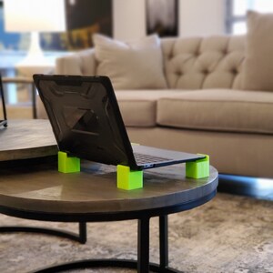 May include: A black laptop computer with a green laptop stand on a gray table. The laptop is open and the screen is facing away from the camera. The table is in a living room setting with a couch in the background.