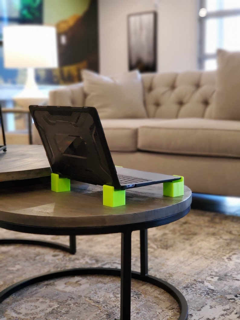 May include: A black laptop computer with a green laptop stand on a gray table. The laptop is open and the screen is facing away from the camera. The table is in a living room setting with a couch in the background.