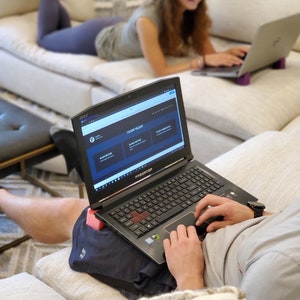 May include: A person using a black laptop computer with a red keyboard and a "PREDATOR" logo on the lid. The laptop is open to a website with the text "TEAM QUIZ" on the screen. The person is sitting on a white couch with a pink cushion.