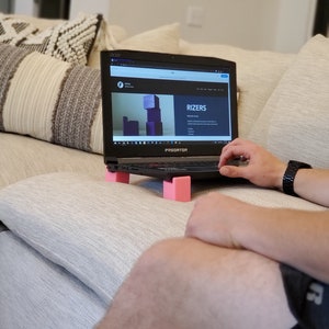 May include: A black laptop computer with a pink laptop stand. The laptop screen shows a website for "Rizers" with a graphic of stacked cubes. The laptop is on a light gray couch.