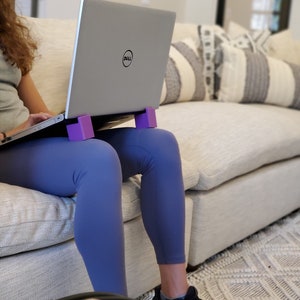 May include: A person sitting on a couch with a laptop computer on their lap. The laptop is on a purple stand. The person is wearing blue leggings and black sneakers.