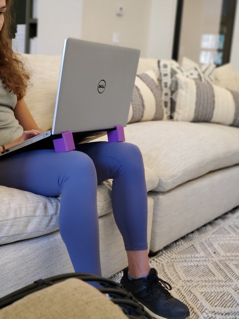 May include: A person sitting on a couch with a laptop computer on their lap. The laptop is on a purple stand. The person is wearing blue leggings and black sneakers.