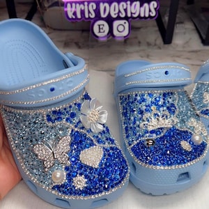 May include: Light blue Crocs adorned with sparkling rhinestones and embellishments. One shoe features a butterfly and flower, the other a crown. The shoes are decorated with various blue and silver gems, creating a glamorous look. The brand name "kris designs" is visible.