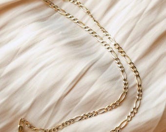 May include: A gold-toned chain necklace featuring a classic Figaro link pattern, which consists of a repeating sequence of three small circular links followed by one elongated oval link. The necklace is secured with a circular spring ring clasp.
