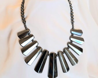 May include: A statement necklace featuring a series of polished, dark grey, metallic-toned beads and elongated, trapezoidal-shaped pendants arranged in a fan-like pattern. The smooth, reflective surfaces of the components create a uniform, monochromatic appearance throughout the entire piece of jewellery.