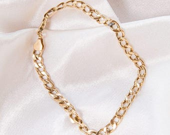 May include: A gold-coloured curb chain bracelet. The bracelet features a series of interlocking oval links. The clasp is a simple, rectangular design. The jewellery is displayed on a white, silky surface.