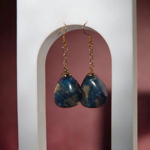 May include: A pair of drop earrings featuring teardrop-shaped pendants with a swirling blue, black, and gold marbled pattern. The pendants are attached to gold-toned metal chains and ear wires, creating a dangling design with a polished finish.