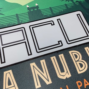 May include: A rectangular white sticker with the letters "ACU" in black, outlined with a black border. The sticker is placed over a background with a green landscape and the words "A Nublar National Park" in gold.