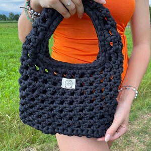 Puede incluir: Una bolsa de mano de crochet negra con una pequeña etiqueta blanca que dice "MADE". La bolsa está siendo sostenida por una persona que lleva una blusa naranja.