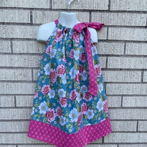 May include: A floral print dress with a halter neckline and a pink bow detail. The dress features a teal background with pink and white flowers, and a pink geometric pattern at the hem. The dress is displayed on a white mannequin.