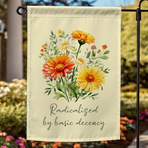 May include: A cream-colored garden flag features a watercolor floral design with orange, yellow, and white gerbera daisies and greenery. The flag has the text "Radicalized by basic decency" in a script font. The flag is hanging on a black metal garden stake.