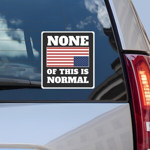 May include: A rectangular sticker with a black border and white text reading "NONE OF THIS IS NORMAL." The sticker features a stylized American flag in red, white, and blue. The sticker is affixed to the rear window of a vehicle.
