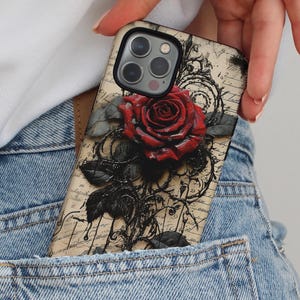 May include: A phone case featuring a detailed red rose with black leaves and vines. The case has a vintage, parchment-like background with script. The case is in a denim pocket.